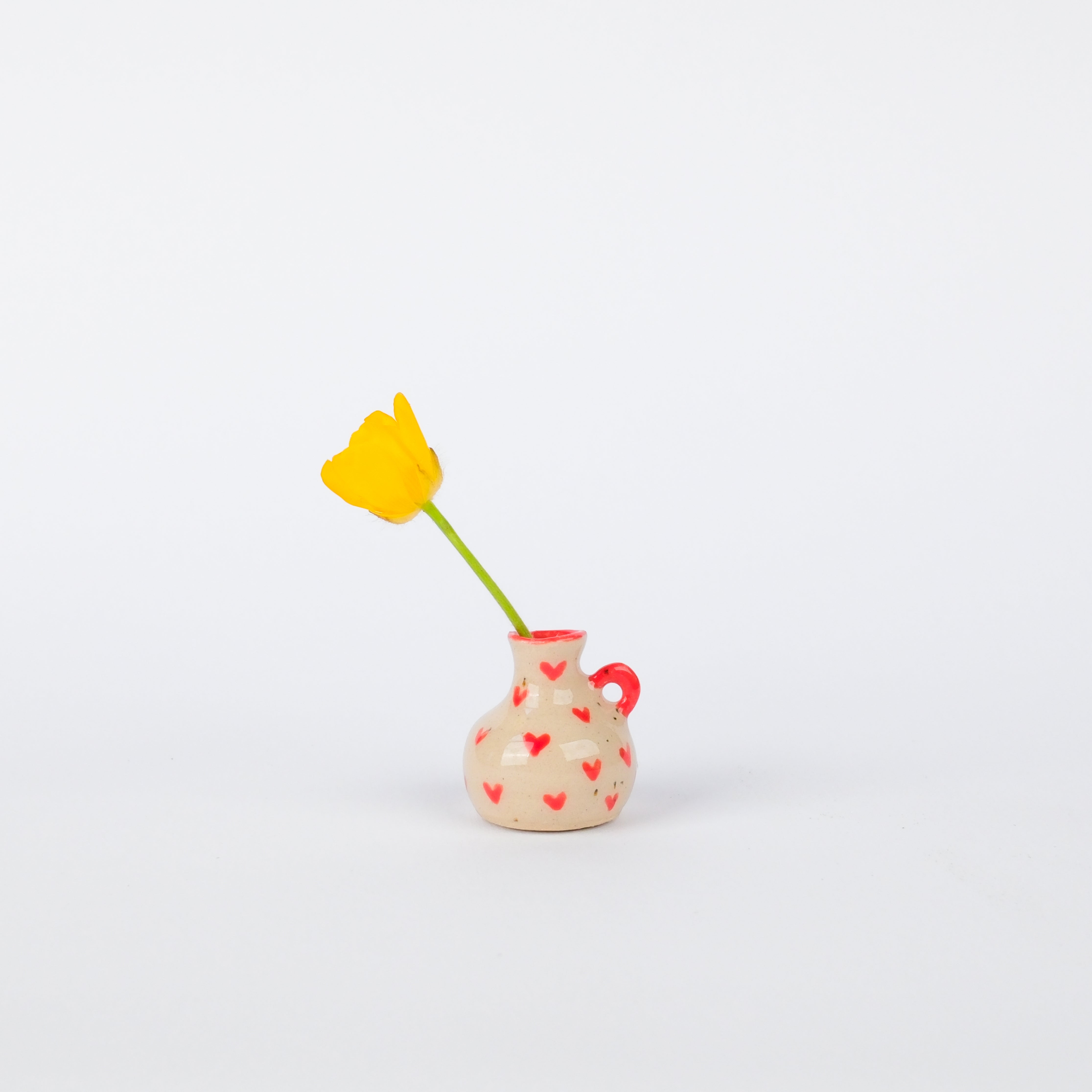 Hand-painted 29mm stoneware vase in speckled cream with tiny red hearts and a small handle, holding a yellow flower.