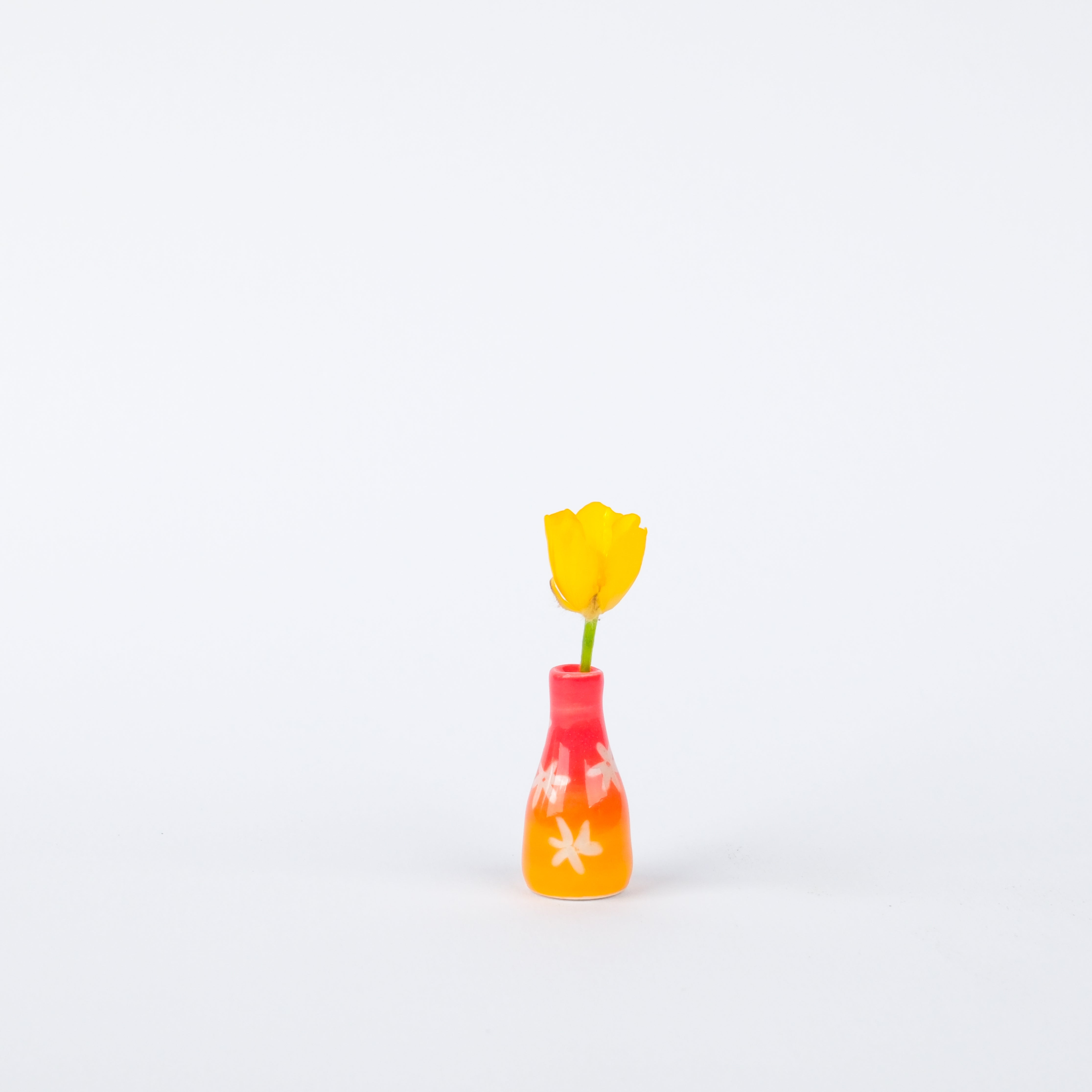 Sunrise Vase 28mm, a tiny handmade stoneware pot with a red-to-orange gradient and white floral carvings holding a yellow flower.