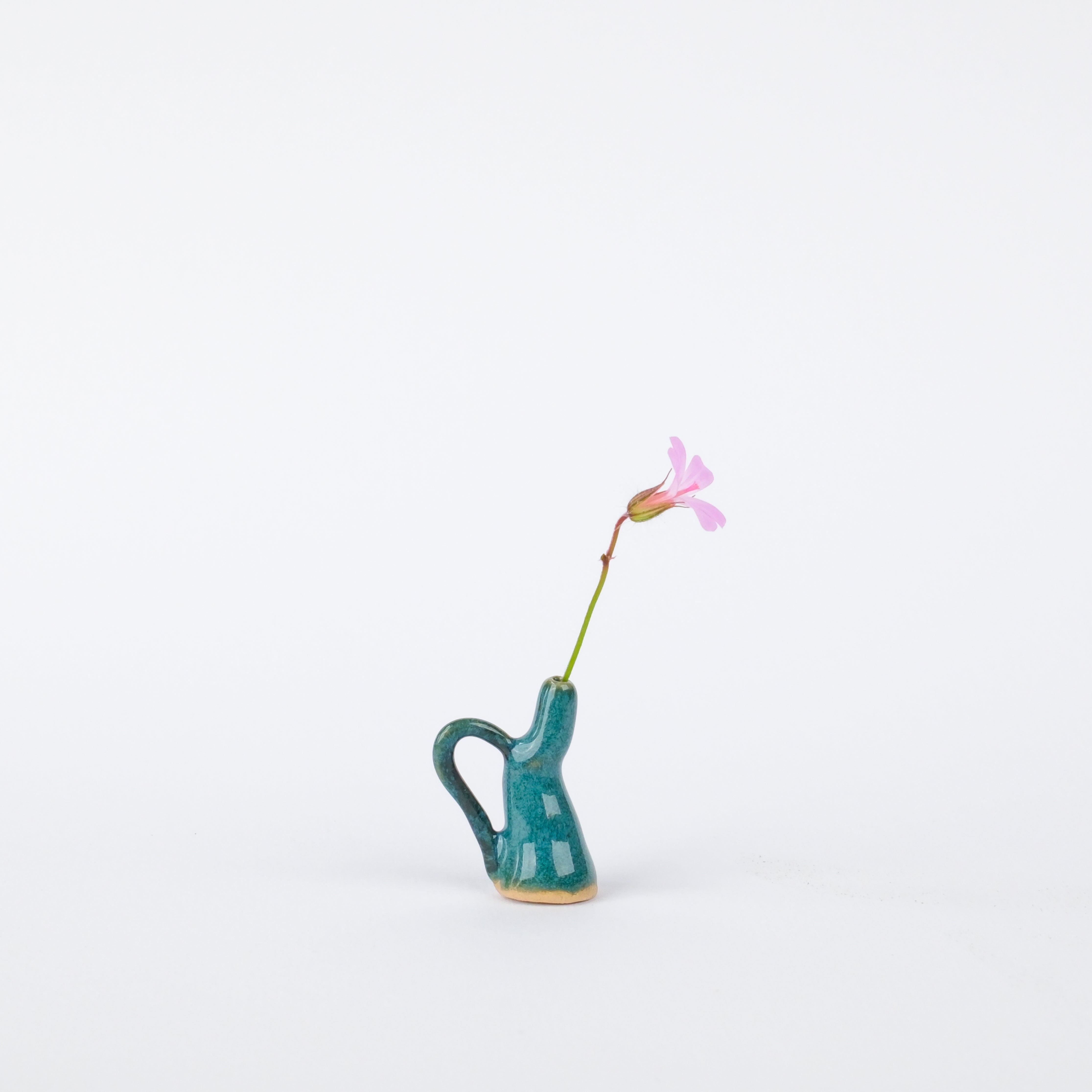 Hand-painted teal stoneware tiny vase with a handle, holding a single small pink flower against a white background.